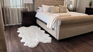 Un dormitorio con una cama con cabecera capitonada, dos mesitas de noche con lámparas, una alfombra mullida blanca y vistas a través de cortinas de encaje.