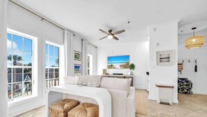 Una sala de estar moderna con un sofá blanco, un ventilador de techo y un ventanal con vistas a palmeras.