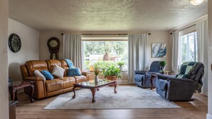 A living room with a brown leather sofa, a glass coffee table, and a grey armchair. There are two clocks on the wall and a window with curtains. - The Rose House: Spacious Retreat w/ Fire Pit Deck/ near expo & Airport/Sleeps 8 (Central Point)
