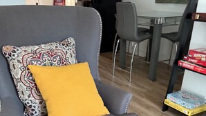 Ein grauer Sessel mit einem gemusterten Kissen und einer senfgelben Decke, ein Esstisch mit Stühlen und ein Bücherregal mit Büchern.