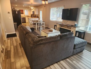 Ein modernes Wohnzimmer mit einem Ecksofa, einem Essbereich mit Tisch und Stühlen, einer Küche im Hintergrund und einem an der Wand montierten Fernseher.
