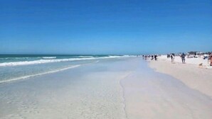 A wide sandy beach with gentle waves, clear skies, and people walking along the shore.