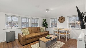 A modern living room with a brown sofa, a wooden coffee table, a dining table with chairs, a large window with blinds, and a clock on the wall.