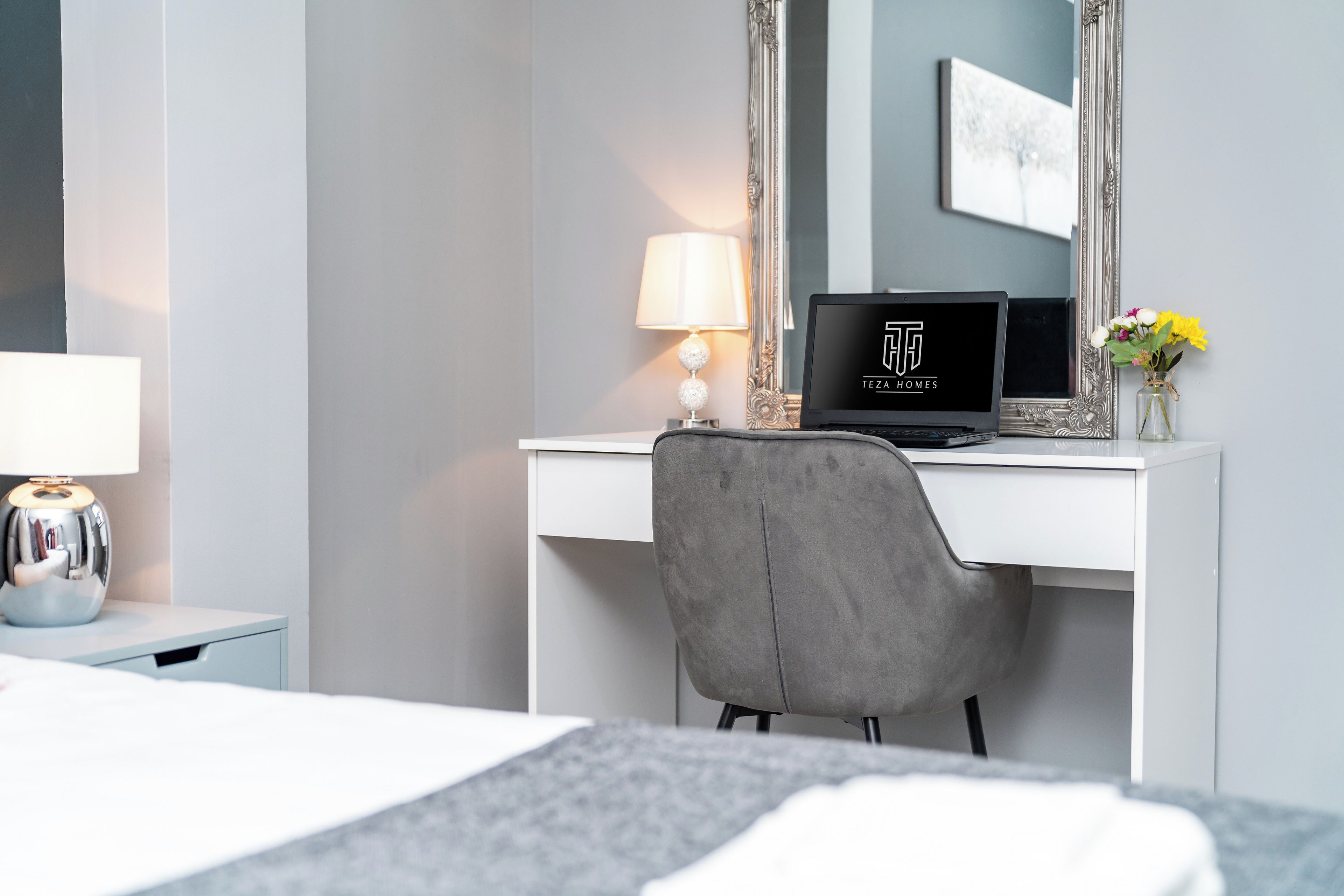 Une chambre d’hôtel avec un bureau blanc, un ordinateur portable, une chaise grise, une table de chevet avec une lampe et un vase rempli de fleurs.