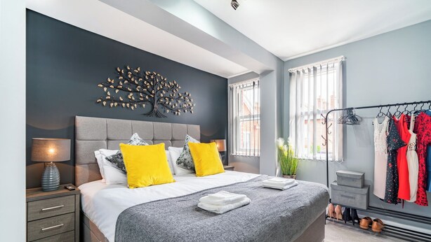 A modern bedroom with a large bed, a tree-shaped wall decoration, a bedside table with a lamp, and a clothing rack with various garments. - Fairfax Close, Amesbury, Wiltshire, SP47ST, United Kingdom (Amesbury)