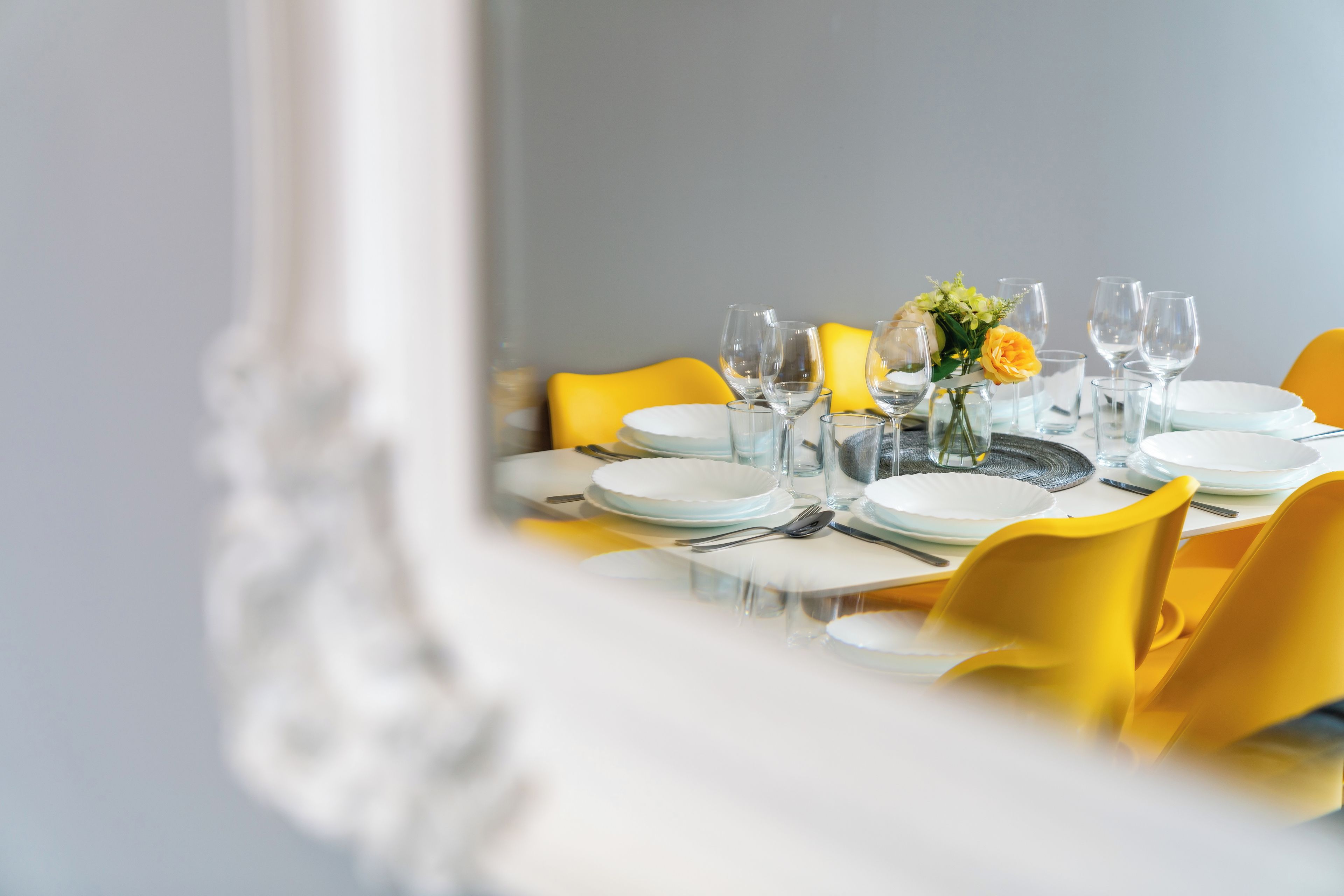 A dining table set with plates, glasses, and cutlery, with yellow chairs and a vase of flowers.