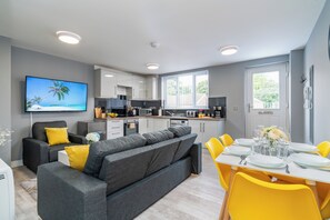 A modern living room with a grey sofa and yellow cushions, a dining area with yellow chairs, a kitchen with white cabinets, and a TV displaying a beach scene. - Fairfax Close, Amesbury, Wiltshire, SP47ST, United Kingdom (Amesbury)