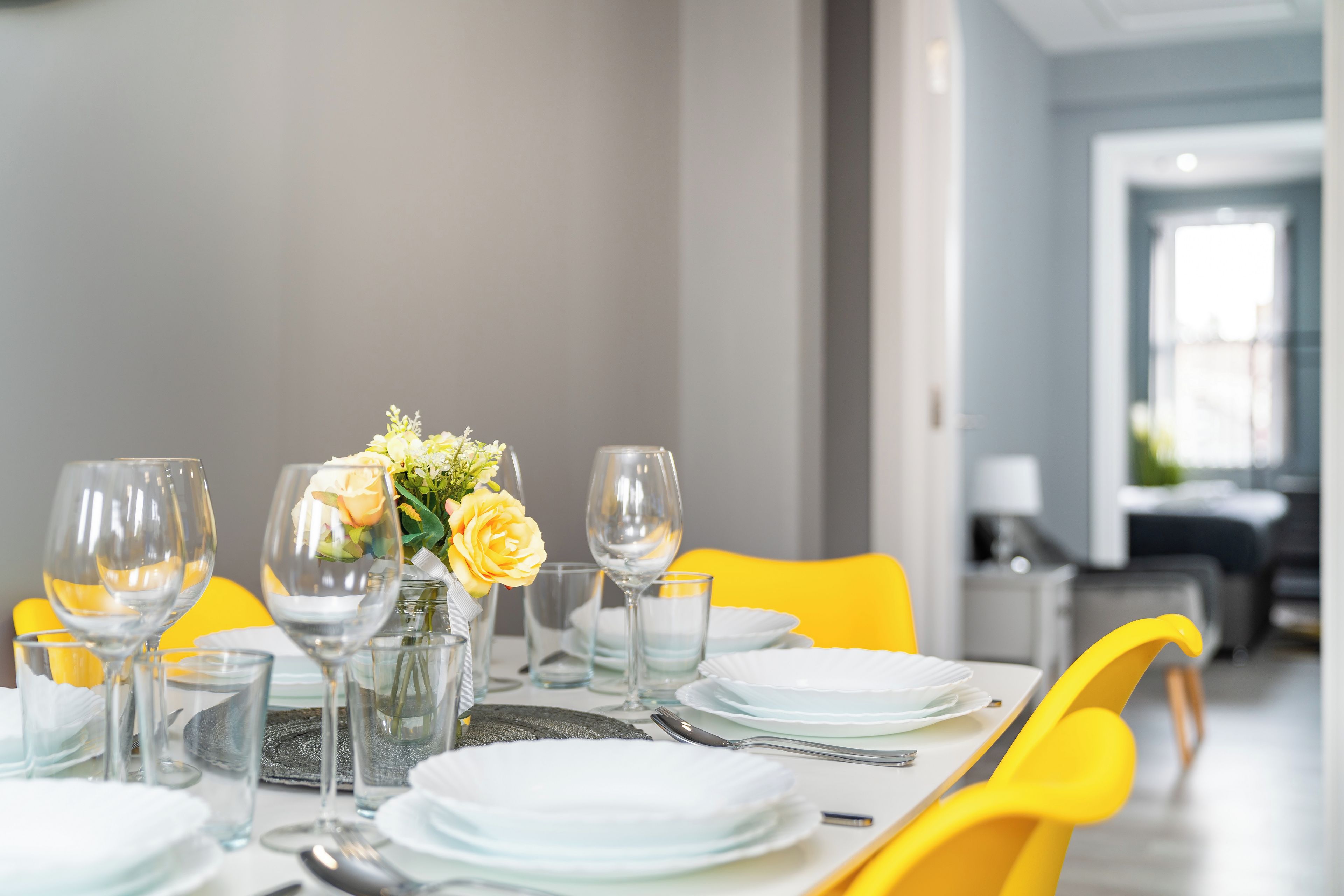 A dining table set with plates, glasses, and cutlery, with a vase of yellow flowers in the center.