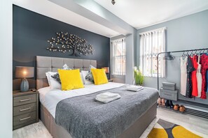 A modern bedroom with a large bed, a decorative wall piece, a window with blinds, and a clothing rack with various garments. - Fairfax Close, Amesbury, Wiltshire, SP47ST, United Kingdom (Amesbury)