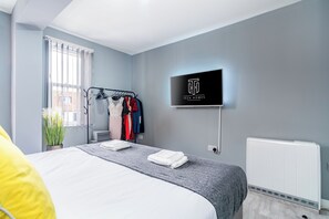 Una habitación de hotel con cama, televisor montado en la pared, radiador y perchero con varias prendas.