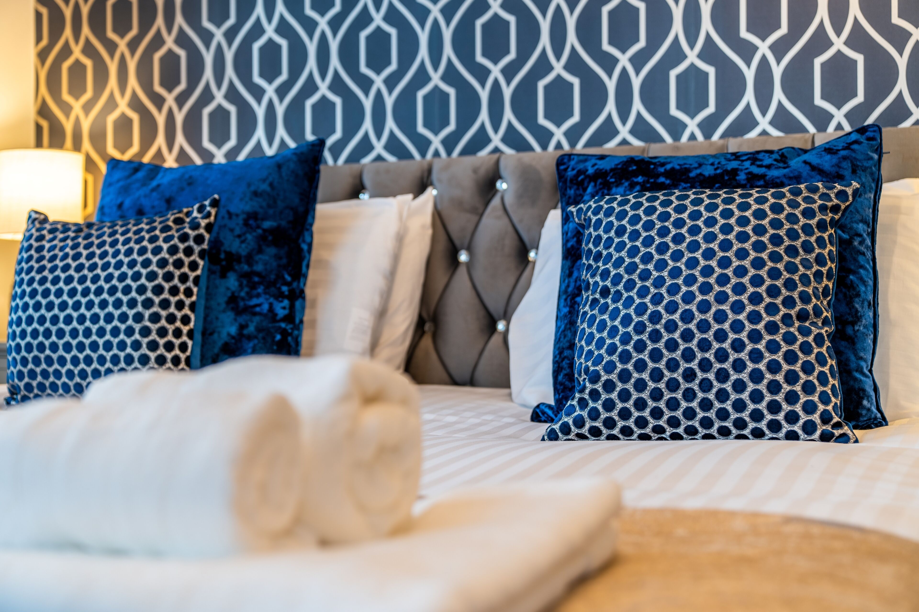A neatly made bed with blue patterned pillows, a tufted headboard, and a geometric patterned wall in the background.