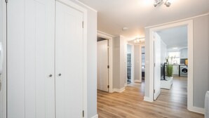 Un couloir au plancher en bois et aux murs blancs, qui mène à un coin cuisine équipé d’un lave-linge et agrémenté d’une plante.