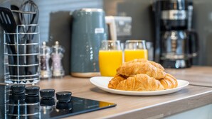 Une table mise pour le petit-déjeuner avec des croissants sur une assiette, une machine à café et un verre de jus d’orange.