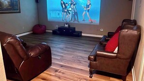 Une salle de cinéma privée dotée d’un grand écran diffusant l’émission « The Mickey Mouse Club », de fauteuils inclinables en cuir et d’un plancher en bois.