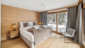 A modern bedroom with a wooden wall, a large bed, a bedside table, a chair, and a view of a snowy landscape. - Chalet Les Rahâs by Mrs Miggins 21 (Grimentz)