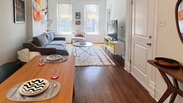 Een moderne woonkamer met een eethoek, houten vloeren, een plafondventilator en een groot raam dat voor natuurlijk licht zorgt.