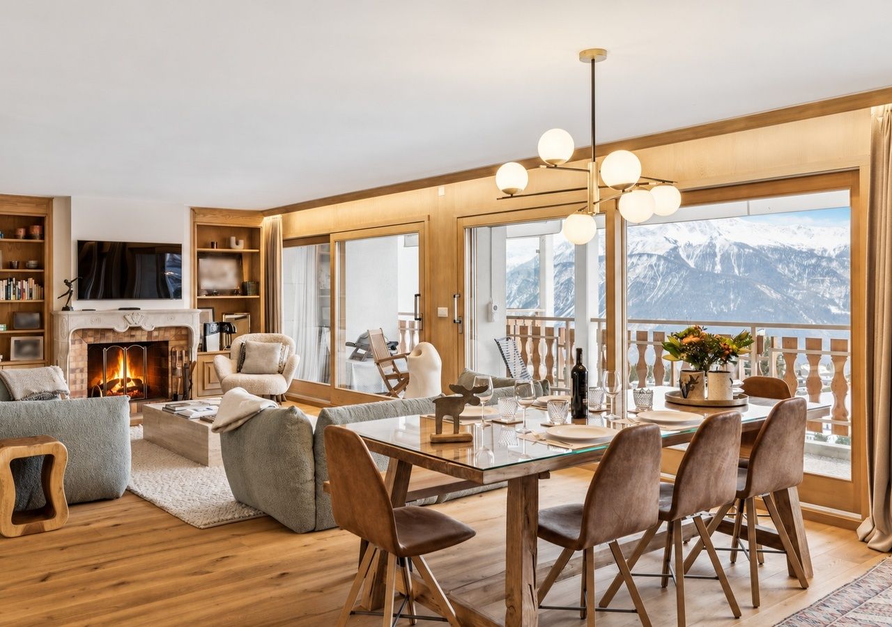 Un salon moderne doté d’une cheminée, d’un coin repas et de grandes fenêtres offrant une vue sur les montagnes.