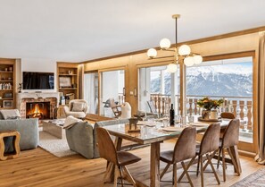 Un salon moderne doté d’une cheminée, d’un coin repas et de grandes fenêtres offrant une vue sur les montagnes.