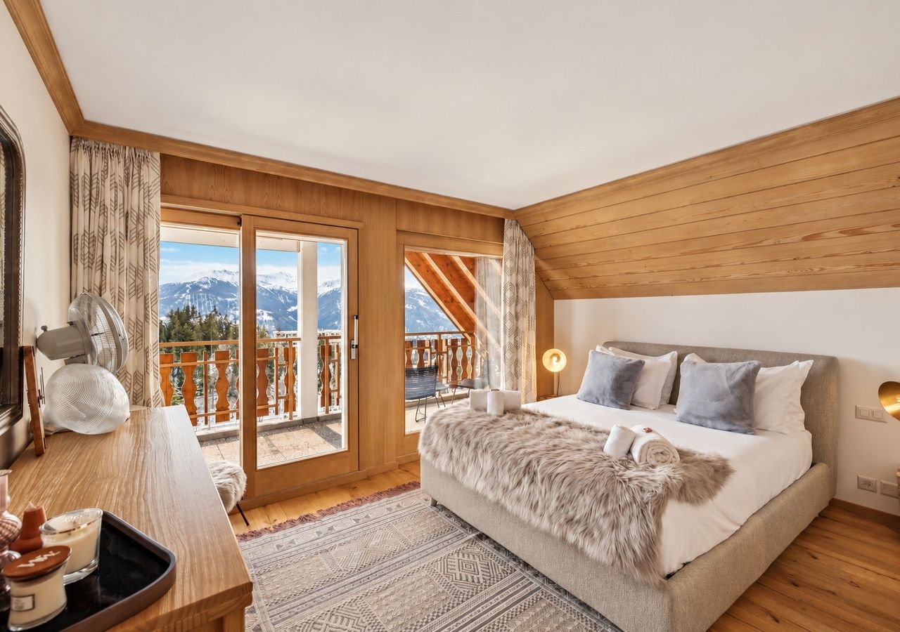 Un dormitorio con una cama grande, techo de madera, vistas a montañas desde un balcón y un escritorio con lámpara.