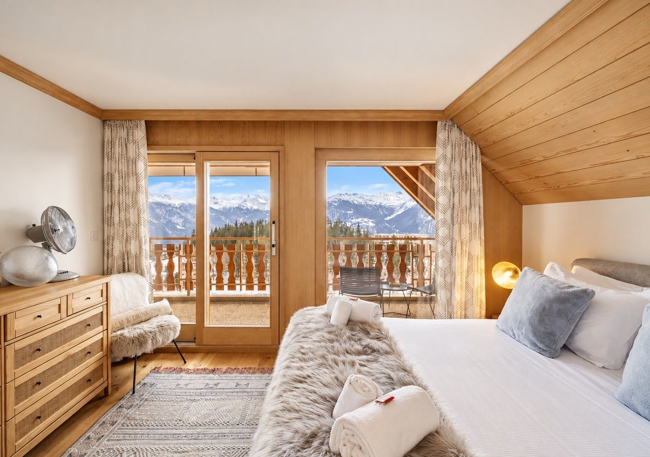A bedroom with a wooden ceiling, a bed with white and blue bedding, a fur throw, a dresser, a fan, and a balcony with a mountain view.