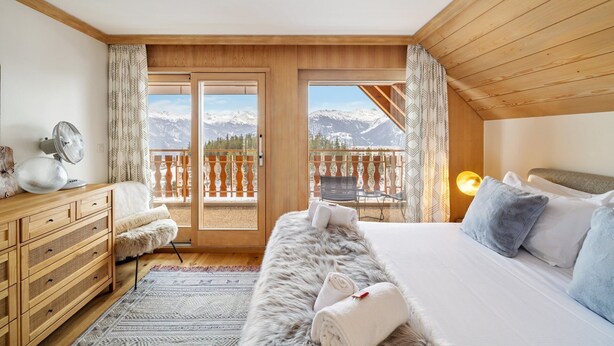 A bedroom with a wooden ceiling, a bed with white and blue pillows, a fluffy chair, a wooden dresser, and a view of snow-capped mountains through a balcony. - Starlight by Mrs Miggins (Crans-Montana)