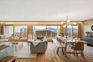 A spacious living room with a wooden floor, a large dining table, and multiple seating options. - Starlight by Mrs Miggins (Crans-Montana)