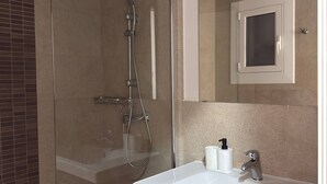 Un baño moderno con ducha envuelta en vidrio, un lavabo blanco con dispensador de jabón negro y un mueble de tocador de madera oscura.