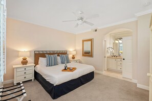 Una camera da letto con un letto, comodini, una ventola a soffitto, uno specchio e un bagno visibile attraverso un arco.