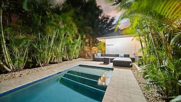 A swimming pool with a tiled deck, surrounded by lush greenery and a seating area with sofas and a table.