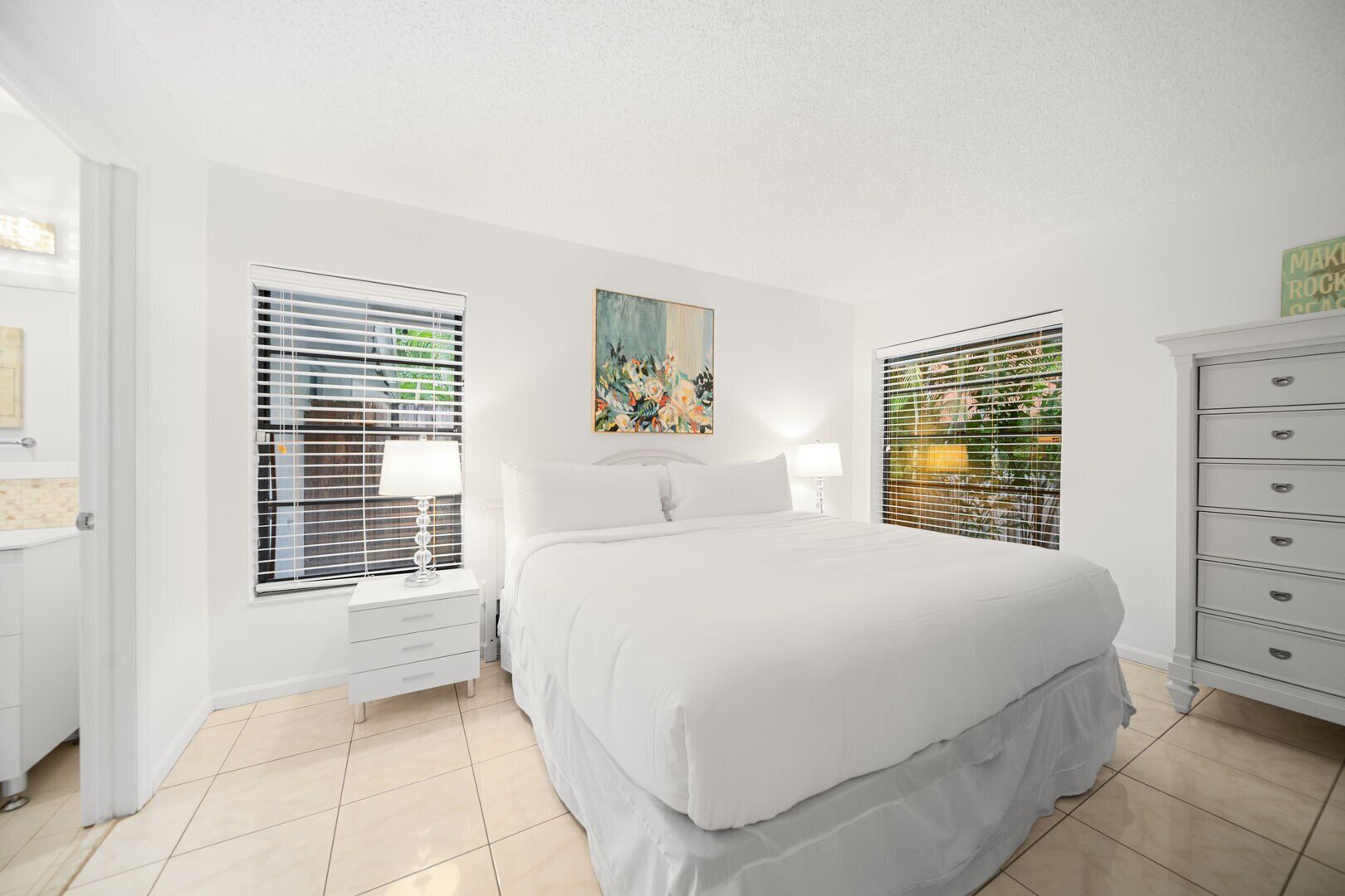 A bedroom with a large bed, two bedside tables, a dresser, and two windows with blinds.