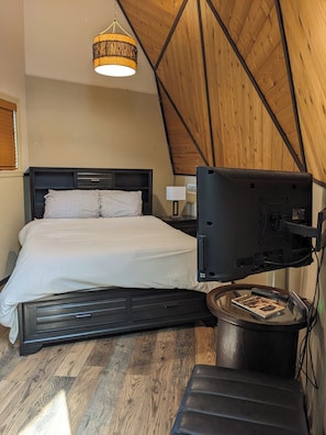 Un dormitorio con una cama, un televisor montado en la pared y una mesita con un libro.