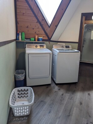 A laundry room with two white washing machines, a shelf with cleaning supplies, a trash bin, and a laundry basket.