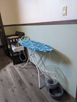 Una habitación con piso de madera, una cama con cubrecama azul estampado, una tabla de planchar y un dispensador de agua.