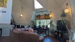A modern living room with a brown leather sofa, a glass coffee table, and a TV stand with a flat-screen TV. There are multiple potted plants, a dining area with a table and chairs, and a kitchen area in the background.
