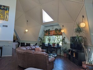 Ein modernes Wohnzimmer mit einem braunen Ledersofa, einem Glastisch und einer TV-Konsole mit einem Flachbildfernseher. Mehrere Blumentöpfe, ein Essbereich mit Tisch und Stühlen sowie eine Küche im Hintergrund.