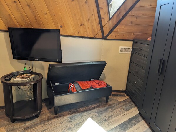 A room with a television mounted on the wall, a small round table with a remote control and a book, a black sofa with a red blanket, and a wardrobe with multiple drawers.