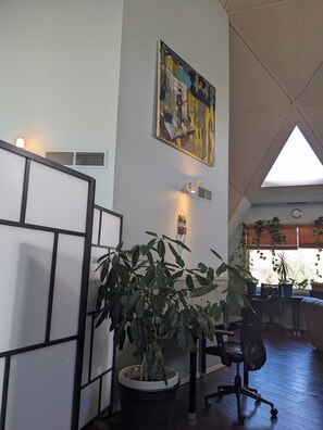 Une pièce avec une grande plante en pot, un tableau accroché au mur et une fenêtre avec des stores.