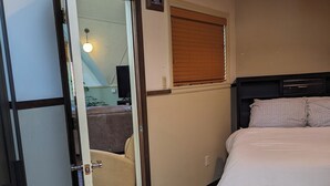 A bedroom with a bed, a door leading to another room, a window with blinds, and a wall-mounted light fixture.