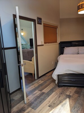 Ein Schlafzimmer mit einem Bett, einer Tür zu einem anderen Raum, einem Fenster mit Jalousien und einer Wandleuchte.