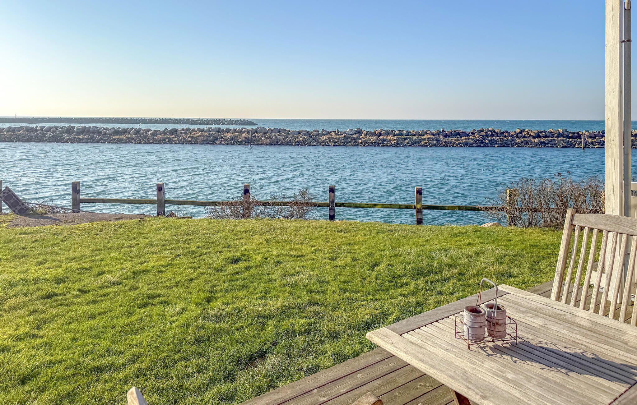 Eine Holzterrasse mit Tisch und zwei Eimern, mit Blick auf ruhiges Wasser und eine felsige Küste.
