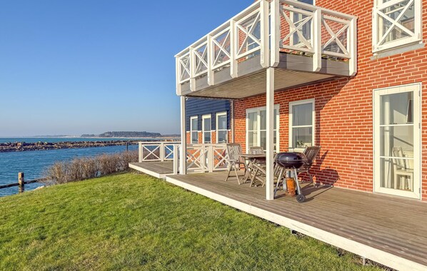 A two-story brick house with a wooden deck, a barbecue grill, and a view of the sea.