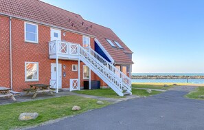 A red brick building with a white staircase leading to a balcony, a paved driveway, and a view of the sea.