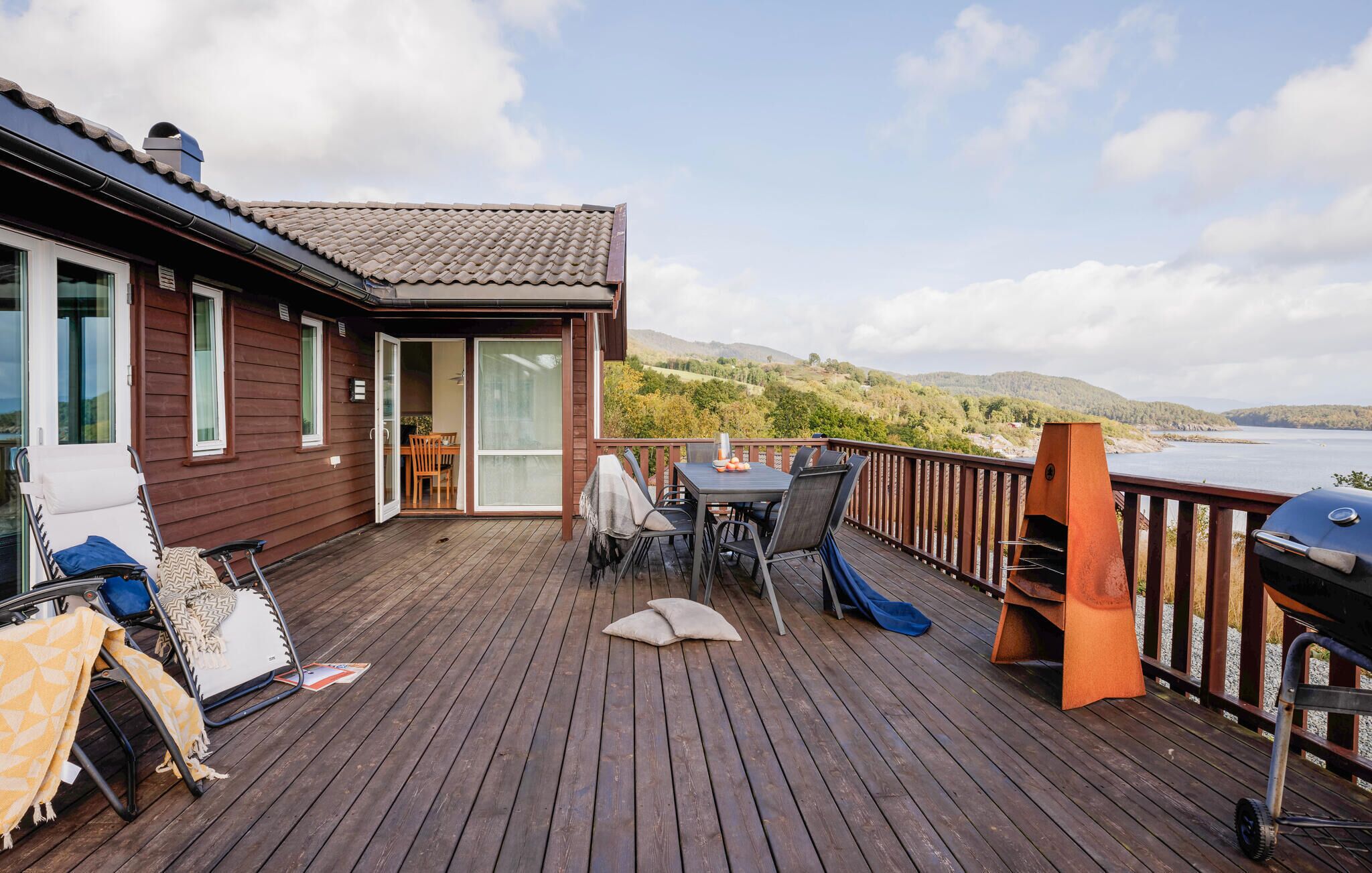 A wooden deck with a table and chairs, a barbecue grill, and a view of the water and hills.