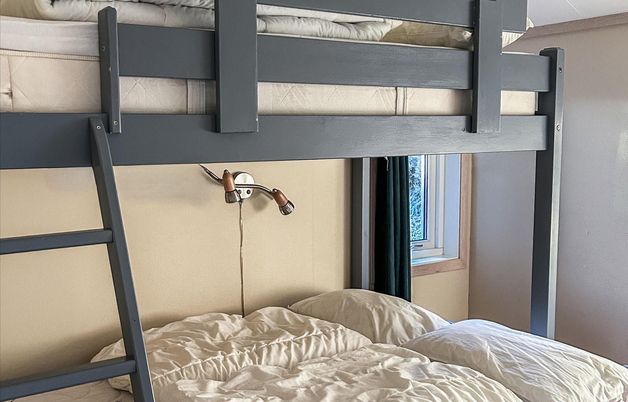 A bunk bed with a ladder, a window with curtains, and a wall-mounted light fixture.