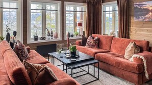 A cozy living room with a large sofa, a coffee table, and a fireplace. There are multiple windows with curtains, and a painting on the wall. - Stunning home in Bjorli with sauna (Bjorli)