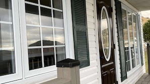 A covered porch with white siding and green shutters, a black trash can, and a wooden railing. - Newfoundland’s Quiet Coast Calls (Harbour Breton)