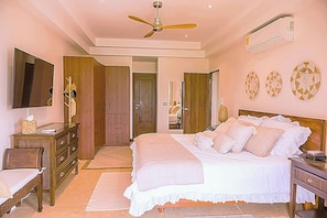 A bedroom with a bed, a ceiling fan, an air conditioner, a dresser, a mirror, and a wall with decorative plates.