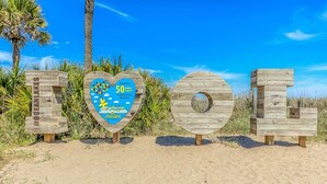 Des lettres en bois formant les mots « JE T’AIME » sont disposées sur une plage, avec un palmier et un ciel dégagé en arrière-plan.