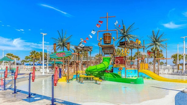 Un parc aquatique avec un toboggan aquatique vert, une zone de jeux avec des jets d’eau et un espace piscine.