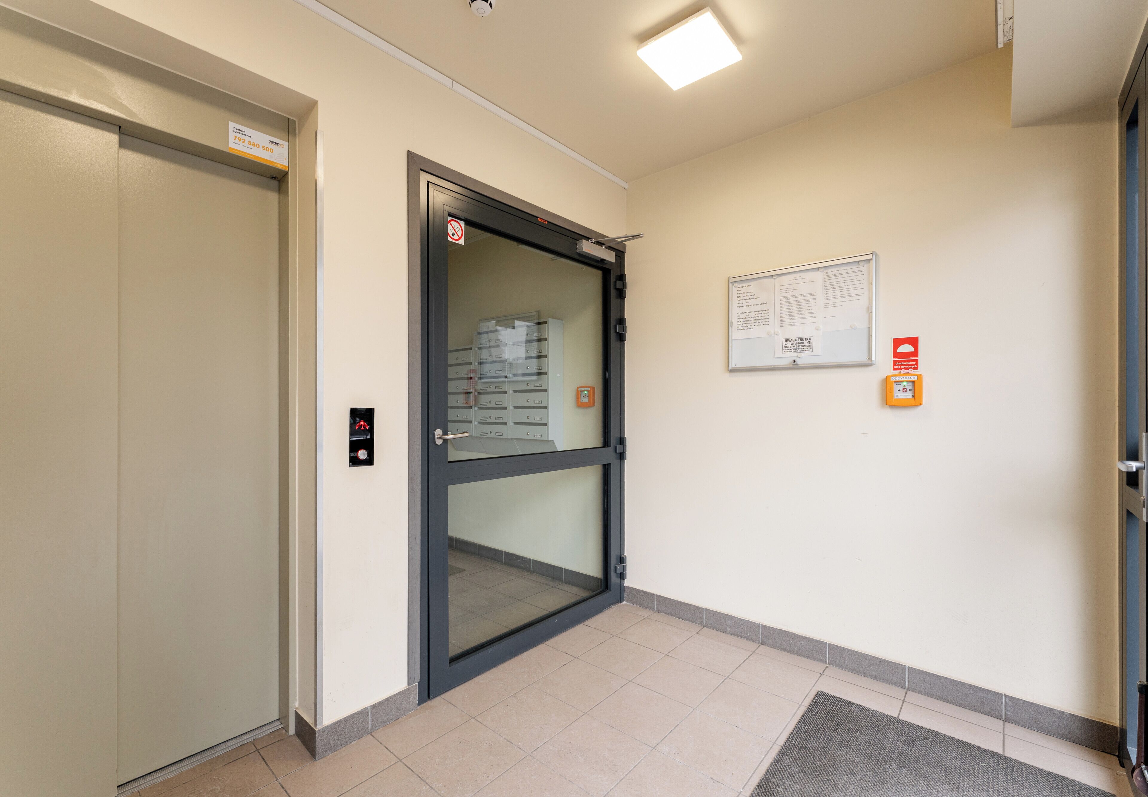 A hotel entrance with a glass door, a fire alarm, and a notice board.